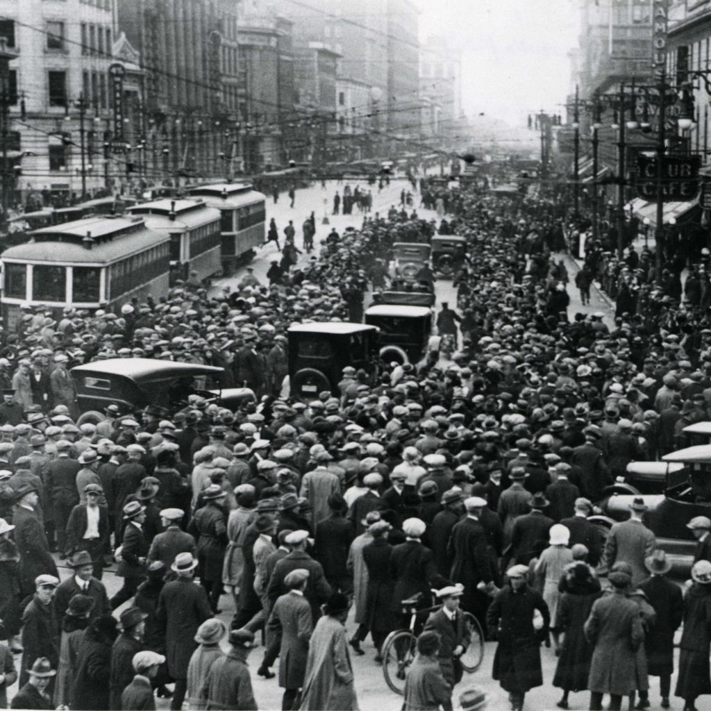 Black and white photograph showing crowds of people in downtown Winnipeg during the Winnipeg General Strike. automobiles and streetcars move between the crowds. Source: University of Manitoba Archives & Special Collections.